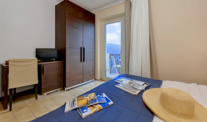 Zimmer mit Bett und Balkon mit Blick auf den Gardasee, Strohhut und Zeitschriften.
